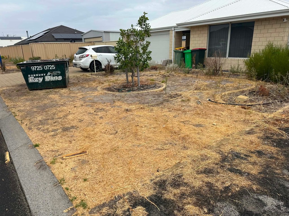 Dry, dead, dirt-covered front yard with a skip bin parked on it.