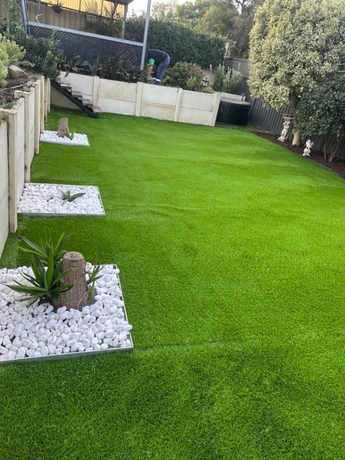 The same backyard transformed with lush green turf, stepping-stone garden islands and white pebble borders.